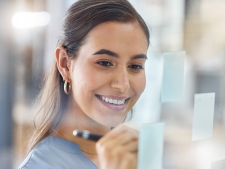 Kvinna ler och skriver på post-it-lappar på en glasvägg i ett kontorslandskap.