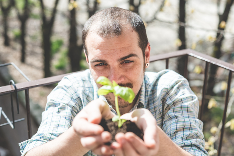 En man håller en liten planta med jord i händerna och ler mot kameran utomhus.
