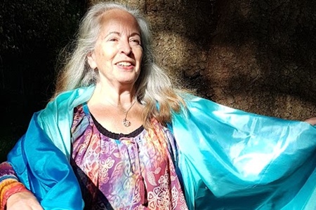 Cirkelledare Elisabeth Hjulström med långt grått hår står barfota utomhus, bär färgglad tunika och blå sjal.