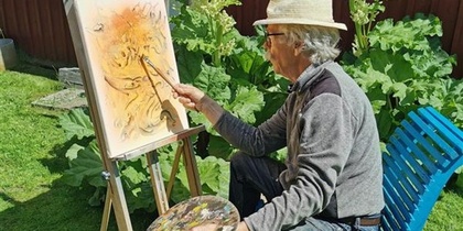 En person målar på en duk utomhus, sittande på en blå stol med en palett i handen.