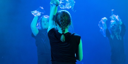 Tre personer i svarta kläder dansar på en scen med blått ljus, en sitter på knä i förgrunden.