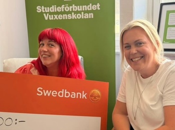 Två personer sitter med en stor orange check från Swedbank på 5000 kronor framför en grön skylt från Vuxenskolan.