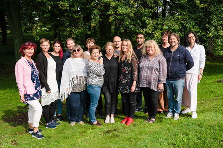 En grupp vuxna står tillsammans utomhus på gräs, leende mot kameran, med träd i bakgrunden.