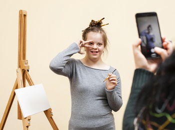 Ung kvinna med penslar i håret poserar framför ett staffli medan någon tar ett foto med mobiltelefon.