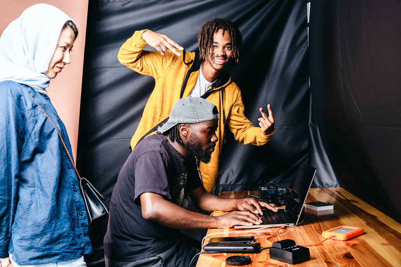 Tre personer arbetar och poserar vid ett träbord med en laptop och teknikprylar, en person gör fredstecken.