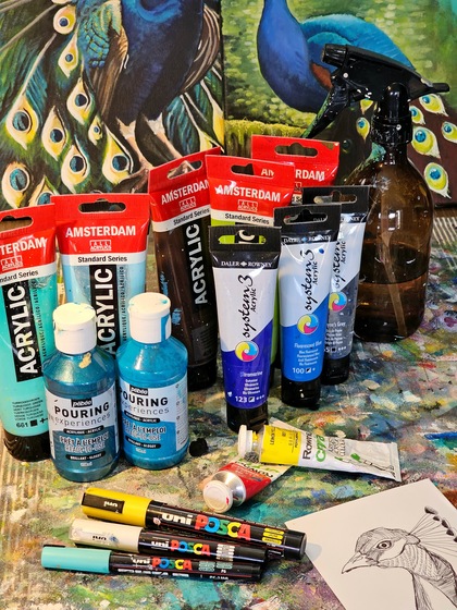 Ett bord med akrylfärger, penslar, markers, en sprayflaska och en teckning av en fågel framför en målning av påfåglar.