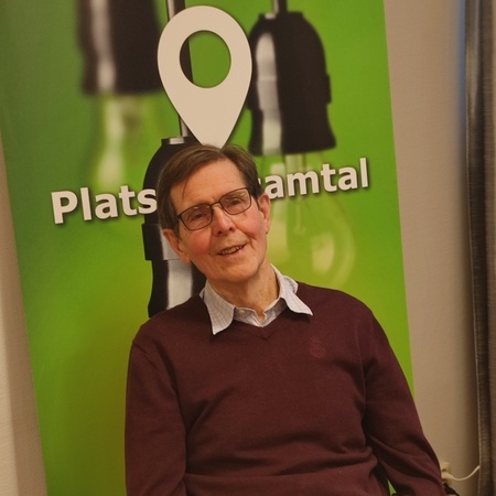 Man med glasögon sitter vid ett bord framför en grön skylt med texten 'Plats för samtal'.