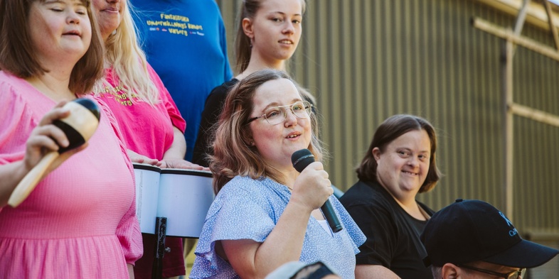 En grupp människor, flera med mikrofon och musikinstrument, står utomhus och sjunger tillsammans.