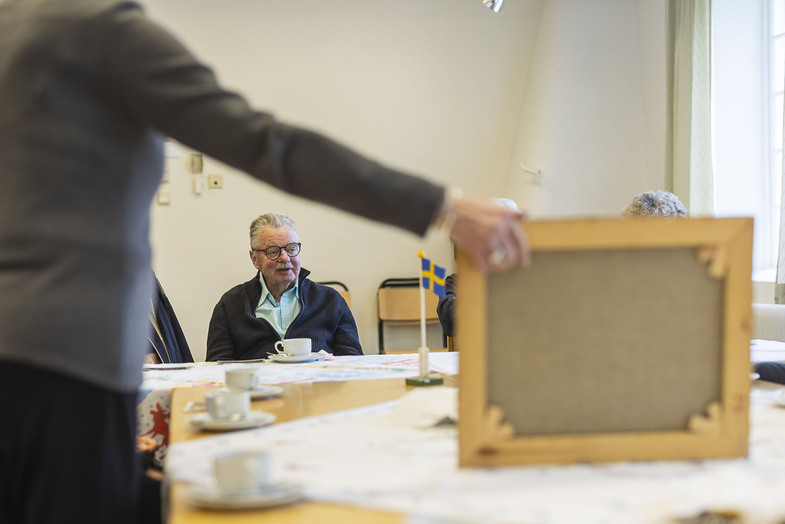 En äldre man sitter vid ett bord med kaffekoppar, medan en person håller upp en tom ram. Svensk flagga syns i bakgrunden.
