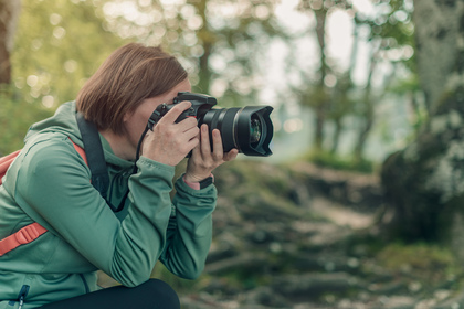 Person med kamera fotograferar i en skogsmiljö, klädd i grön jacka och ryggsäck.