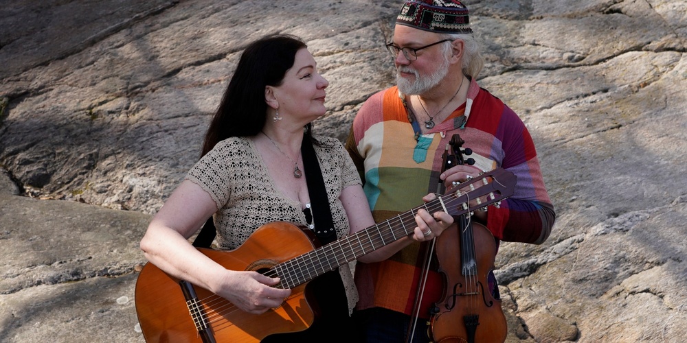 Två personer spelar gitarr och fiol utomhus mot en klippbakgrund.