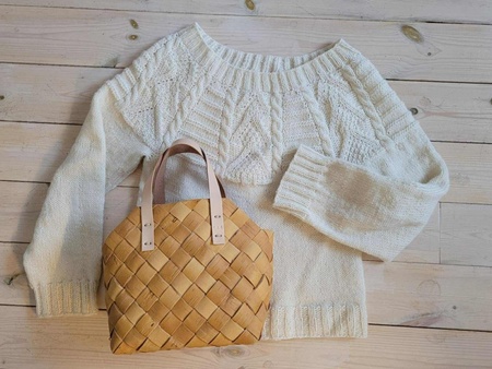 En vit stickad tröja och en flätad korgväska ligger på ett ljust trägolv.