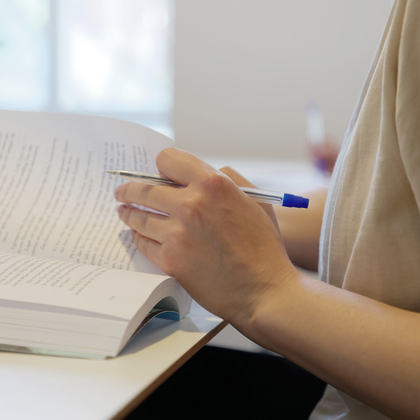 En person sitter vid ett bord och läser en bok, håller en penna i handen.