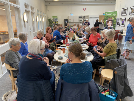 En grupp äldre kvinnor sitter runt bord och handarbetar i en ljus samlingslokal.