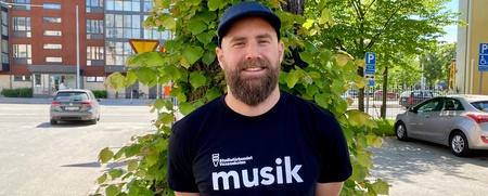 Man med skägg och keps står framför ett träd på en solig gata, bär svart t-shirt med texten 'musik'.