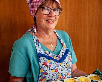 Kvinna med glasögon, förkläde och sjalett håller en plåt med nybakade pajer framför en trävägg.
