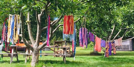 Färgglada tygband hänger mellan träd i en grönskande trädgård med bord och skylt i bakgrunden.