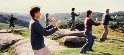 En grupp människor utövar tai chi utomhus på en naturskön plats med kullar och grönska.