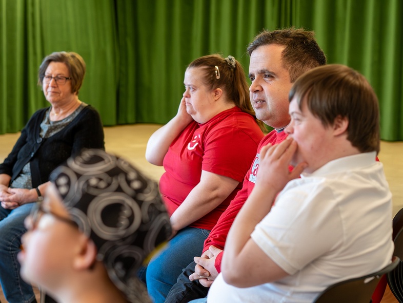 En grupp personer sitter i en cirkel i ett rum med grön gardin i bakgrunden.