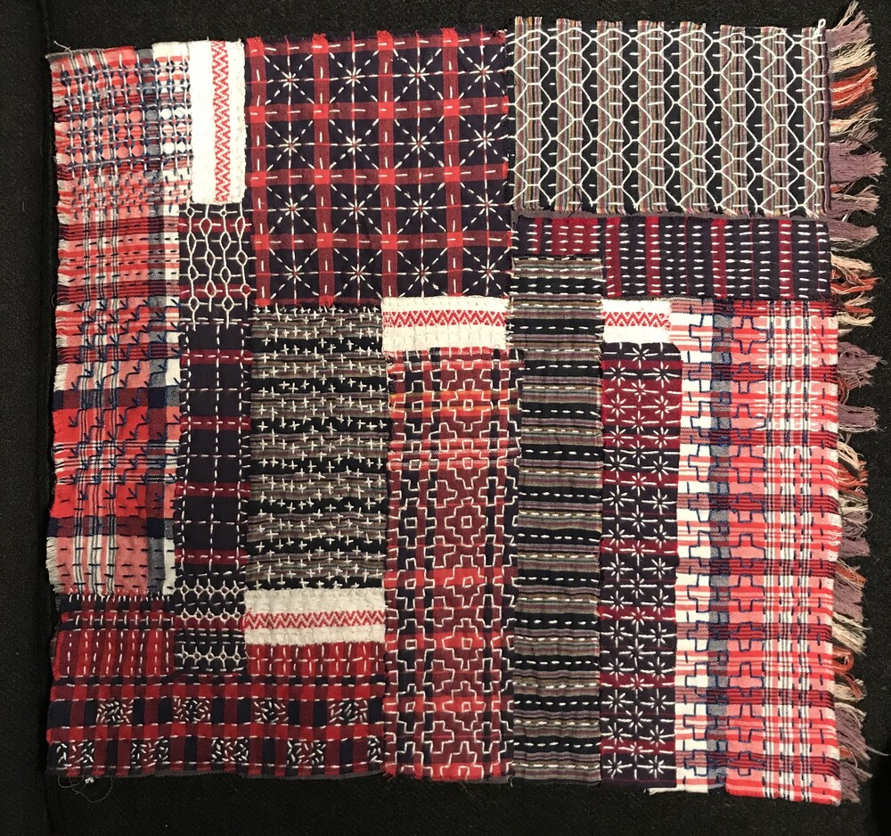 En vävd filt med olika geometriska mönster i rött, blått, vitt och svart, lagd platt mot en mörk bakgrund.