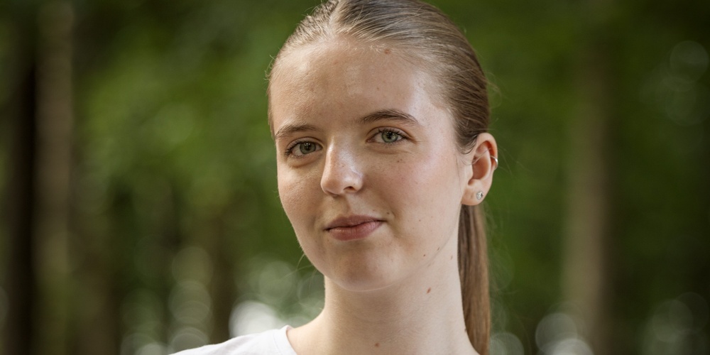 Person med vitt t-shirt står utomhus i en grön skogsmiljö.