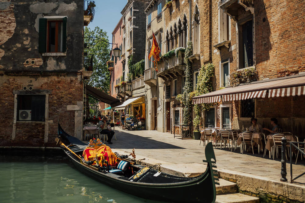 En gondol ligger vid en kanal i Venedig, omgiven av gamla byggnader och ett café med uteservering.