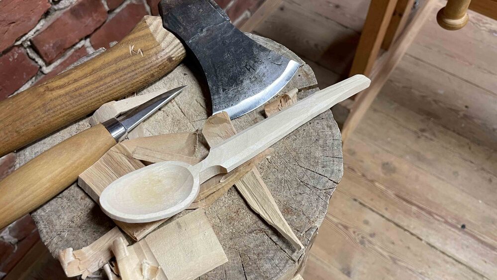 En träslev under tillverkning ligger på en trästubbe med en yxa och en kniv bredvid, omgiven av träspån.