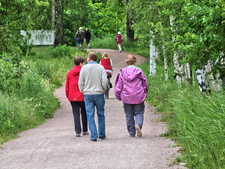 Flera personer promenerar på en grusväg omgiven av gröna träd och gräs en sommardag.