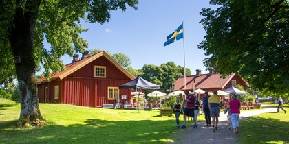 Folk promenerar mot röda trähus med svensk flagga, soligt väder och grönskande omgivning.