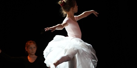 En ung ballerina i vit tyllkjol dansar på scenen med armarna utsträckta, belyst av scenljus.