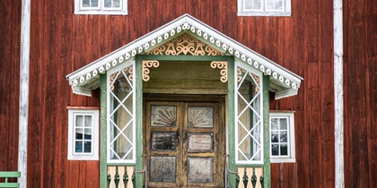 Entré till ett rött trähus med snickarglädje, gröna detaljer och dubbeldörrar i trä.