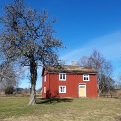 Rött trähus med vita fönster står på en gräsmatta under klarblå himmel, omgiven av träd utan löv.