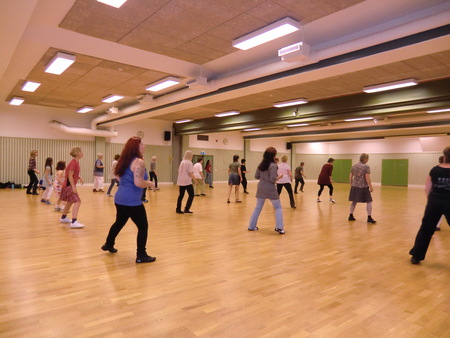 En grupp människor dansar tillsammans i en stor ljus sal med trägolv och högt i tak.