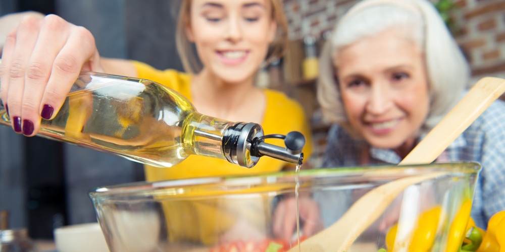 Två kvinnor lagar mat tillsammans och häller olja i en salladsskål med grönsaker.