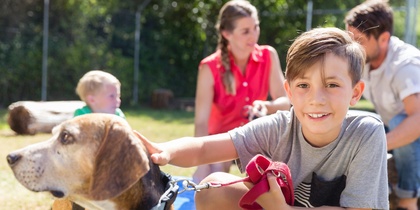 En pojke klappar en hund utomhus medan en familj sitter i bakgrunden på en solig dag.