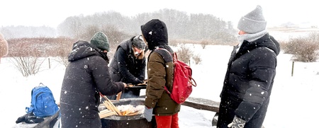 Fyra personer står runt en eld utomhus i ett snöigt vinterlandskap, klädda i vinterkläder.