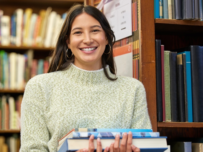 Ung kvinna med långt mörkt hår håller böcker och ler i ett bibliotek.