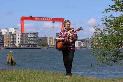 Person spelar gitarr på gräs vid vattnet med Eriksbergs kran och stad i bakgrunden.