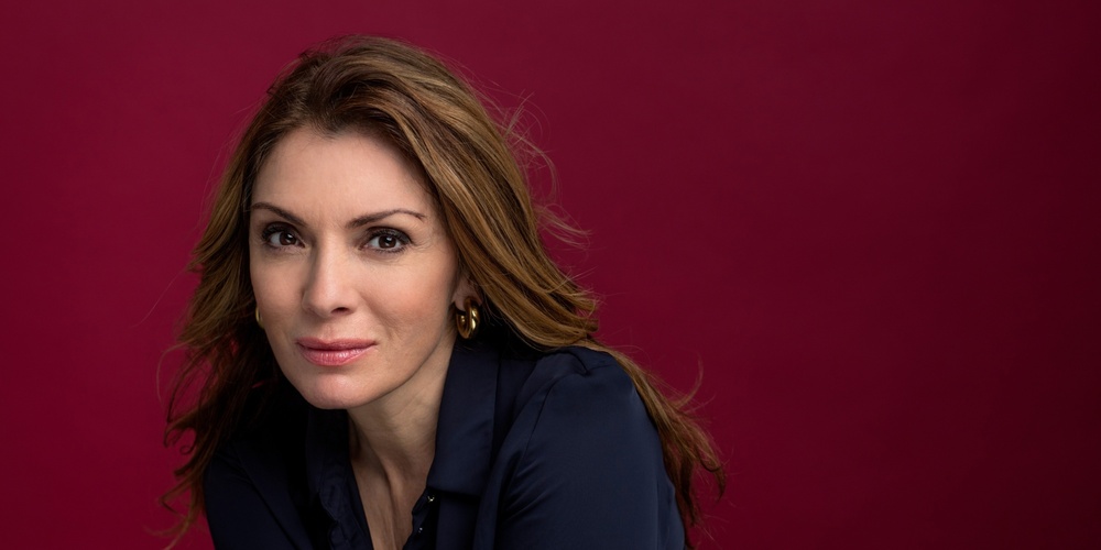 Person med långt brunt hår och mörkblå skjorta mot vinröd bakgrund.