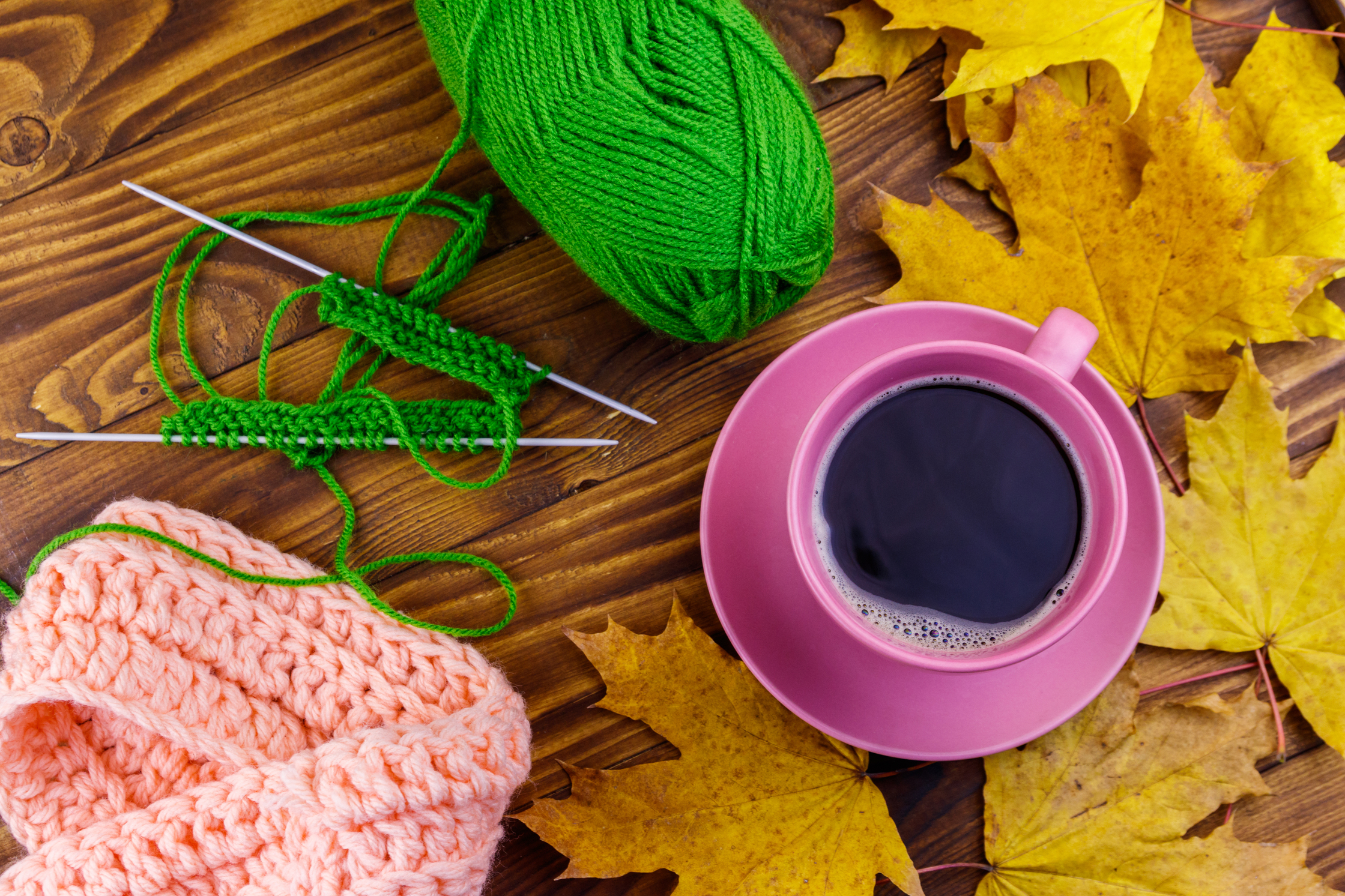 Stickning med grönt garn, rosa stickat plagg, rosa kaffekopp och gula höstlöv på ett träbord.