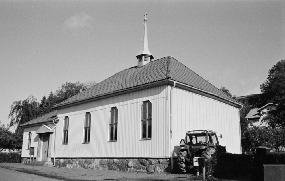En vit träkyrka med brant tak och ett litet torn, en traktor står parkerad bredvid byggnaden.