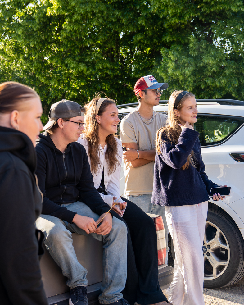 Fem unga personer står och sitter utomhus vid bilar, de ser glada ut och pratar med varandra.