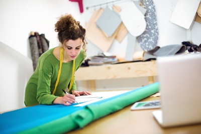 Kvinna i grön tröja ritar mönster på tyg i en syateljé med måttband runt halsen.