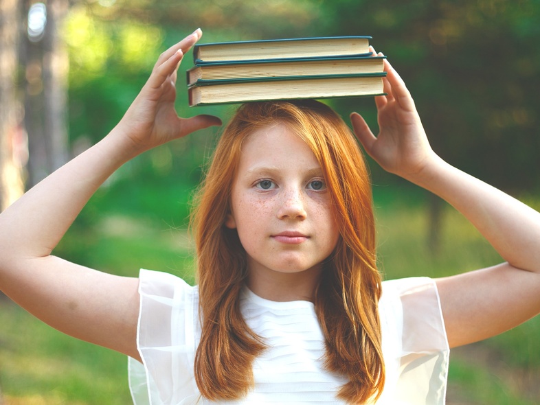 Flicka i vit klänning balanserar tre böcker på huvudet utomhus i en grön skogsmiljö.
