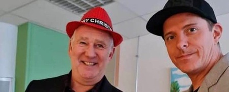 Två män poserar tillsammans inomhus, en med röd hatt och en med svart keps, båda klädda i kavaj.