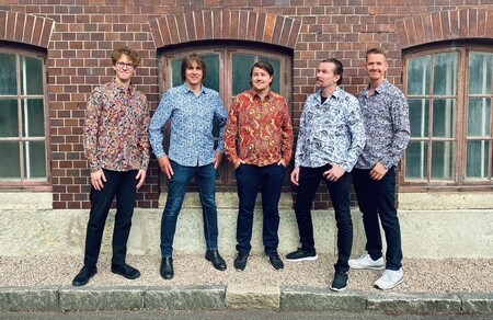 Fem män i mönstrade skjortor står framför en tegelvägg med fönster.