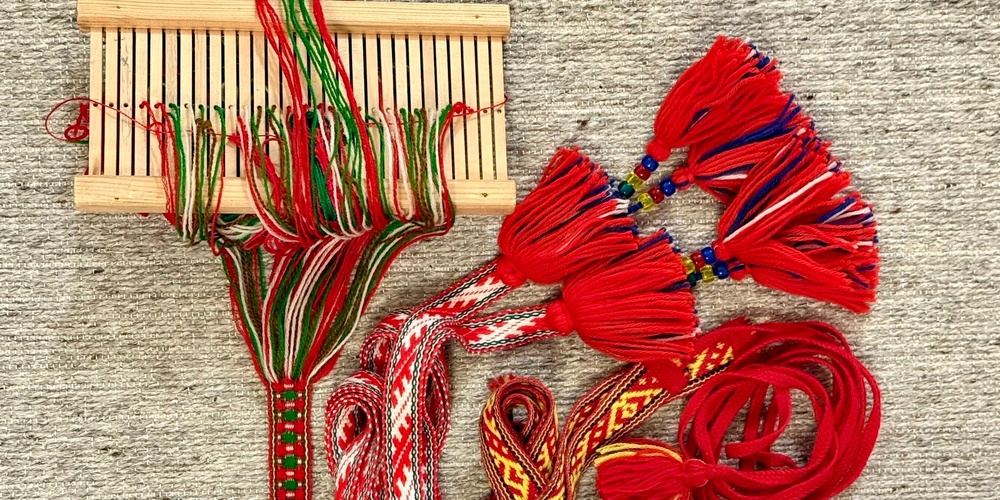 Handvävda band i rött, grönt och vitt med tofsar och pärlor, samt en enkel vävram på en ljus matta.
