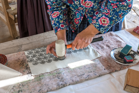 En person trycker mönster på tyg med hjälp av en stämpel och hammare vid ett bord.