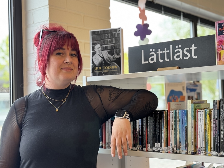 Person med rött hår står vid en bokhylla med skylten 'Lättläst' i ett bibliotek.