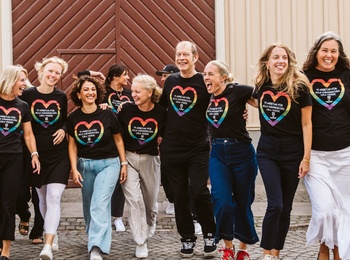 En grupp människor går tillsammans på en kullerstensgata framför en träbyggnad, alla bär svarta t-shirts med regnbågshjärtan.